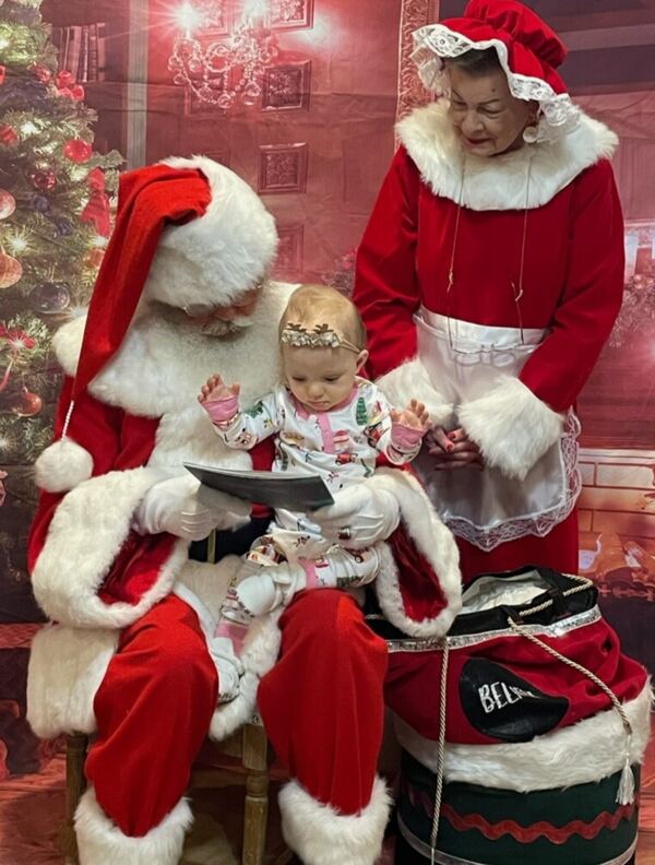 Baby on Santa's lap reading a book, Mrs. Claus looking on