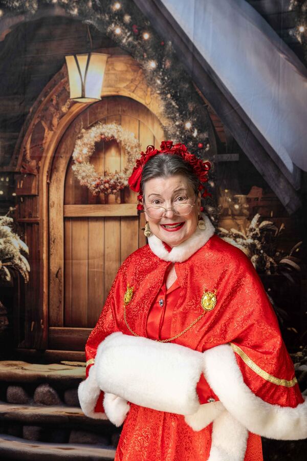 Mrs. Claus Nancy in a red cape and fur muff at a storybook cottage door