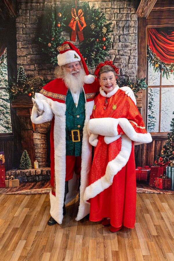 Santa Truette in a long coat and green vest beside Mrs. Claus Nancy in a Christmas parlor