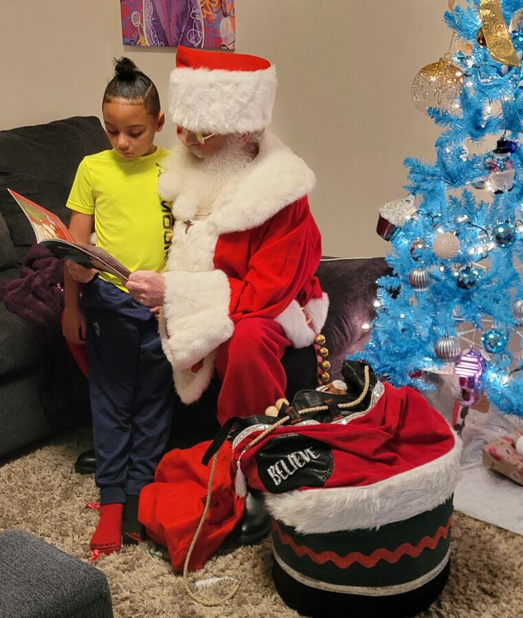 Santa reading a Christmas book with a young girl beside a blue tree